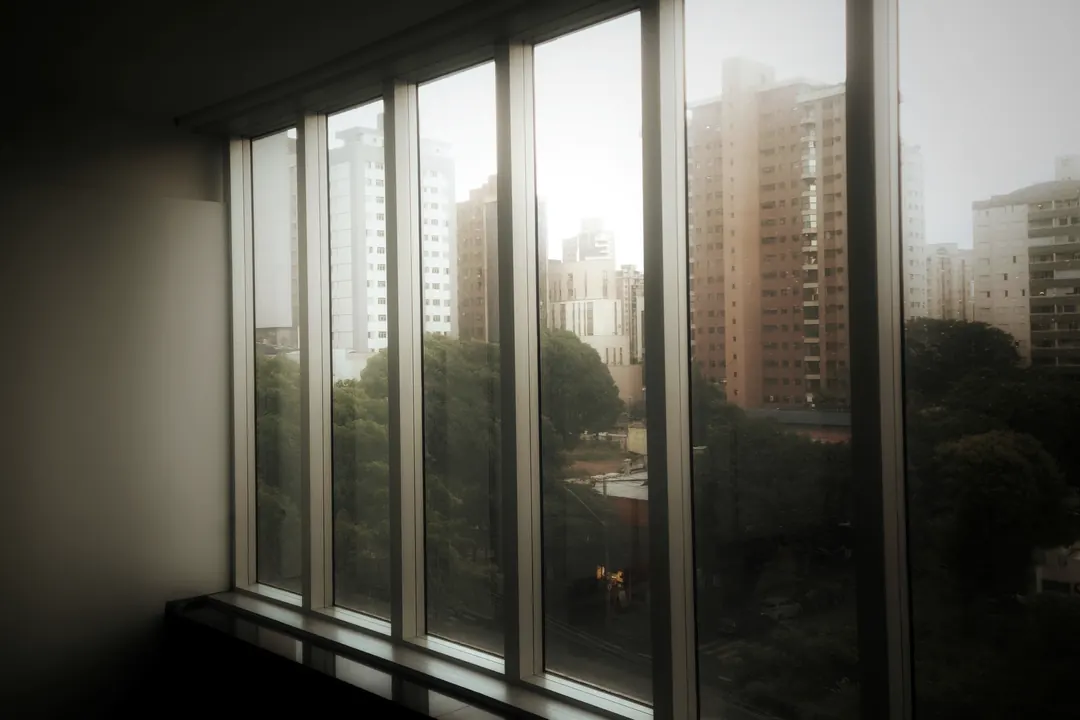 Interior escuro com uma janela longa de perfis finos aberta para árvores e prédios ao entardecer